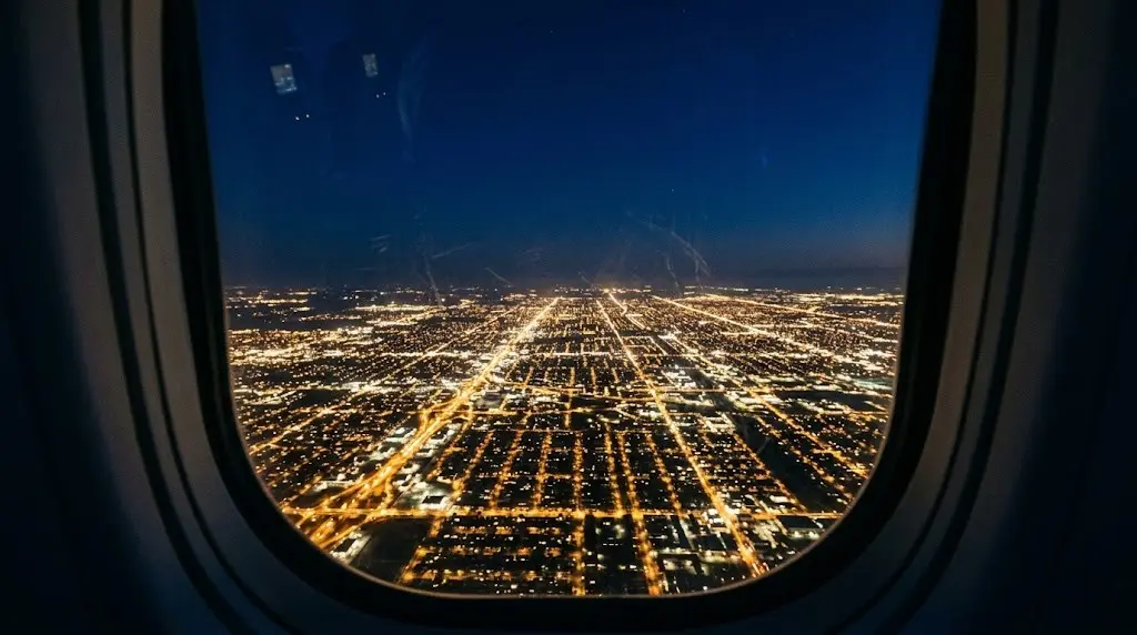 View from airplane window at night showing a glowing city grid far below in darkness
